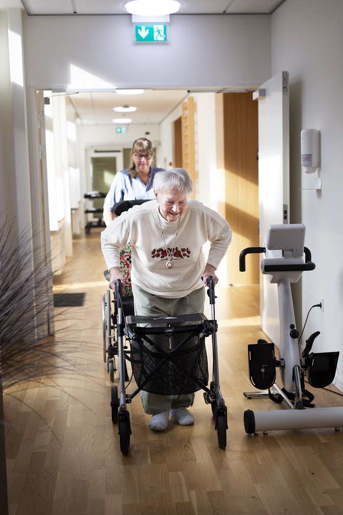 Ældre dame går ned ad en gang. Der er en SOSU-assistent, som følger efter og hjælper hende.