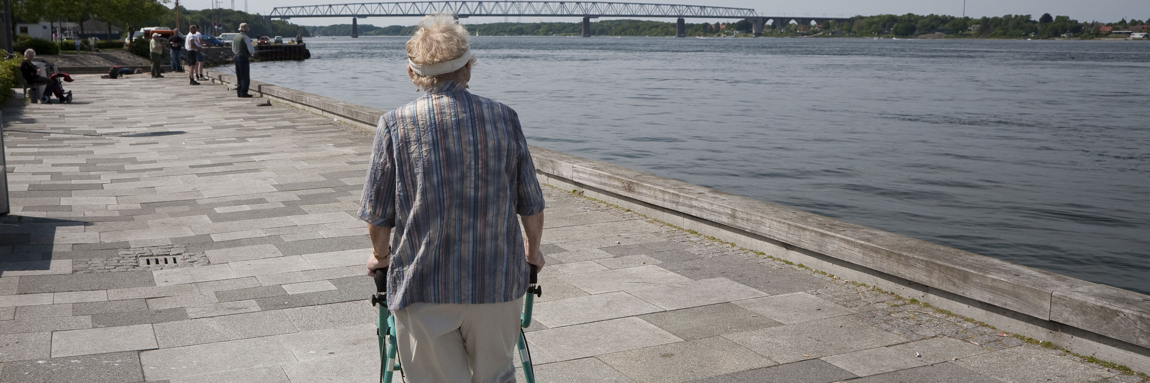 Ældre person går langs en flod. Personen støtter sig op ad sin rollator.