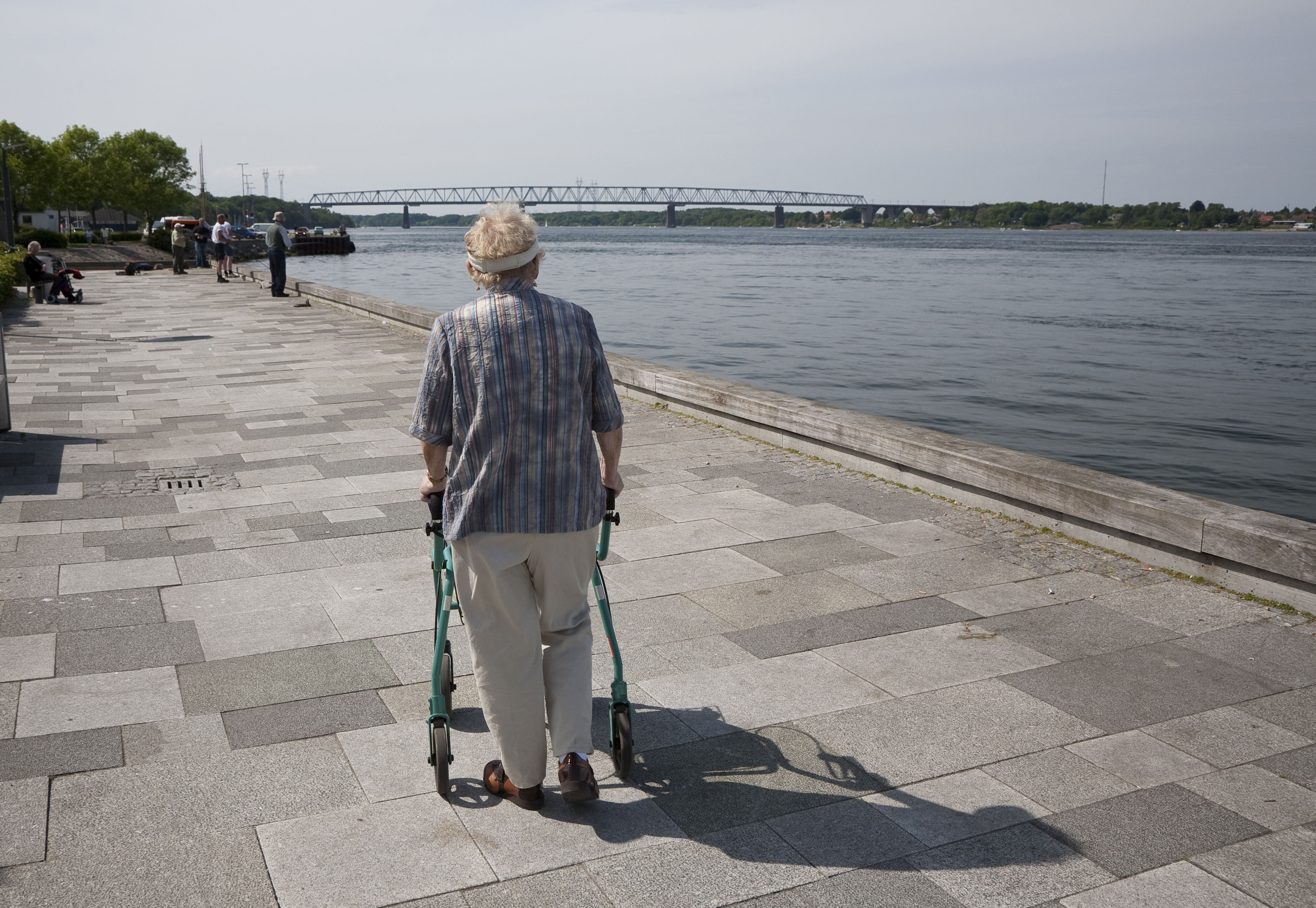 Ældre person går langs en flod. Personen støtter sig op ad sin rollator.