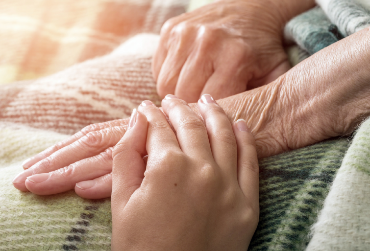 En yngre kvinde holder en ældre borger i hånden. Deres hænder ligger på et tæppe på den ældre persons skød.
