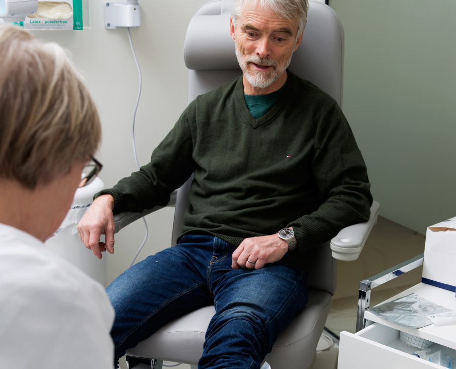 Mand er ved lægen. Han sidder på en stol. I forgrunden sidder en kvindelig sygeplejerske.